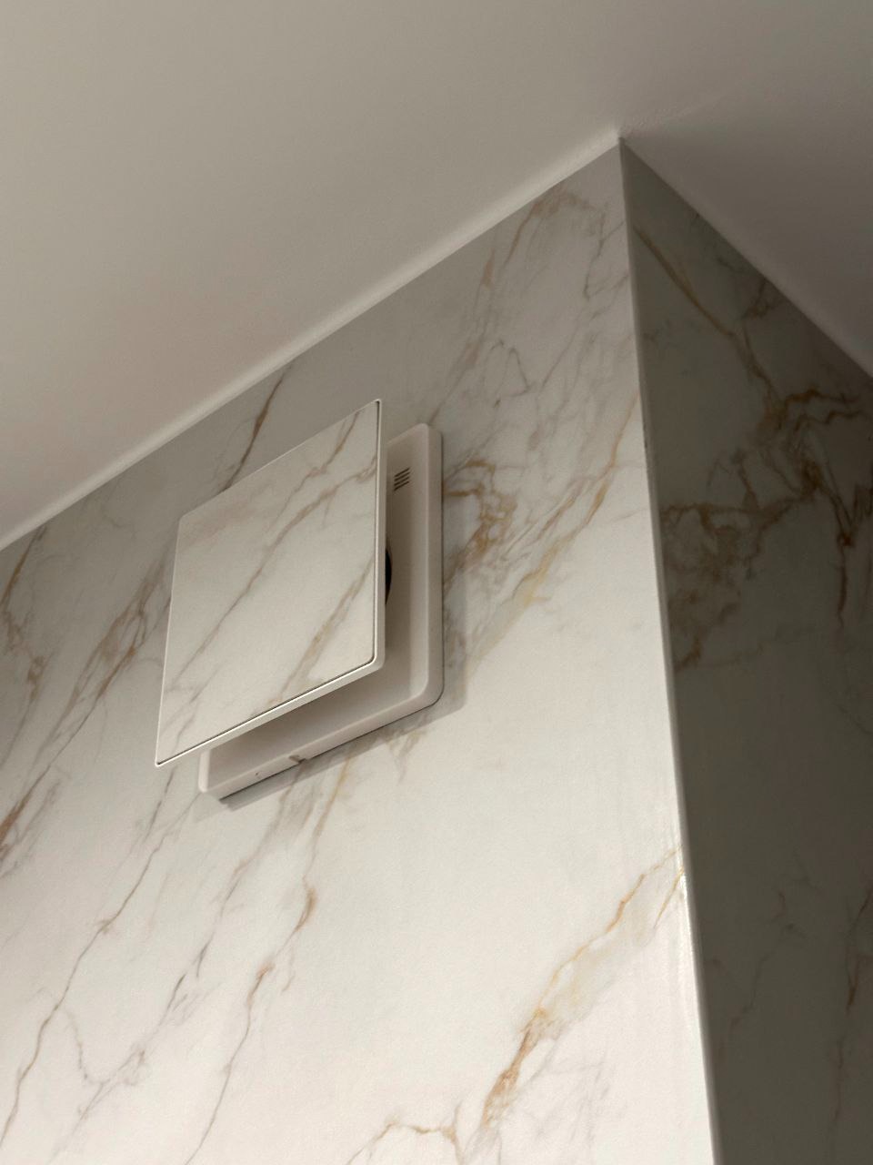 Biały, kwadratowy wentylator z marmurowym wzorem na ścianie wykończonej płytami imitującymi marmur. Ujęcie z dołu, widoczny fragment sufitu.