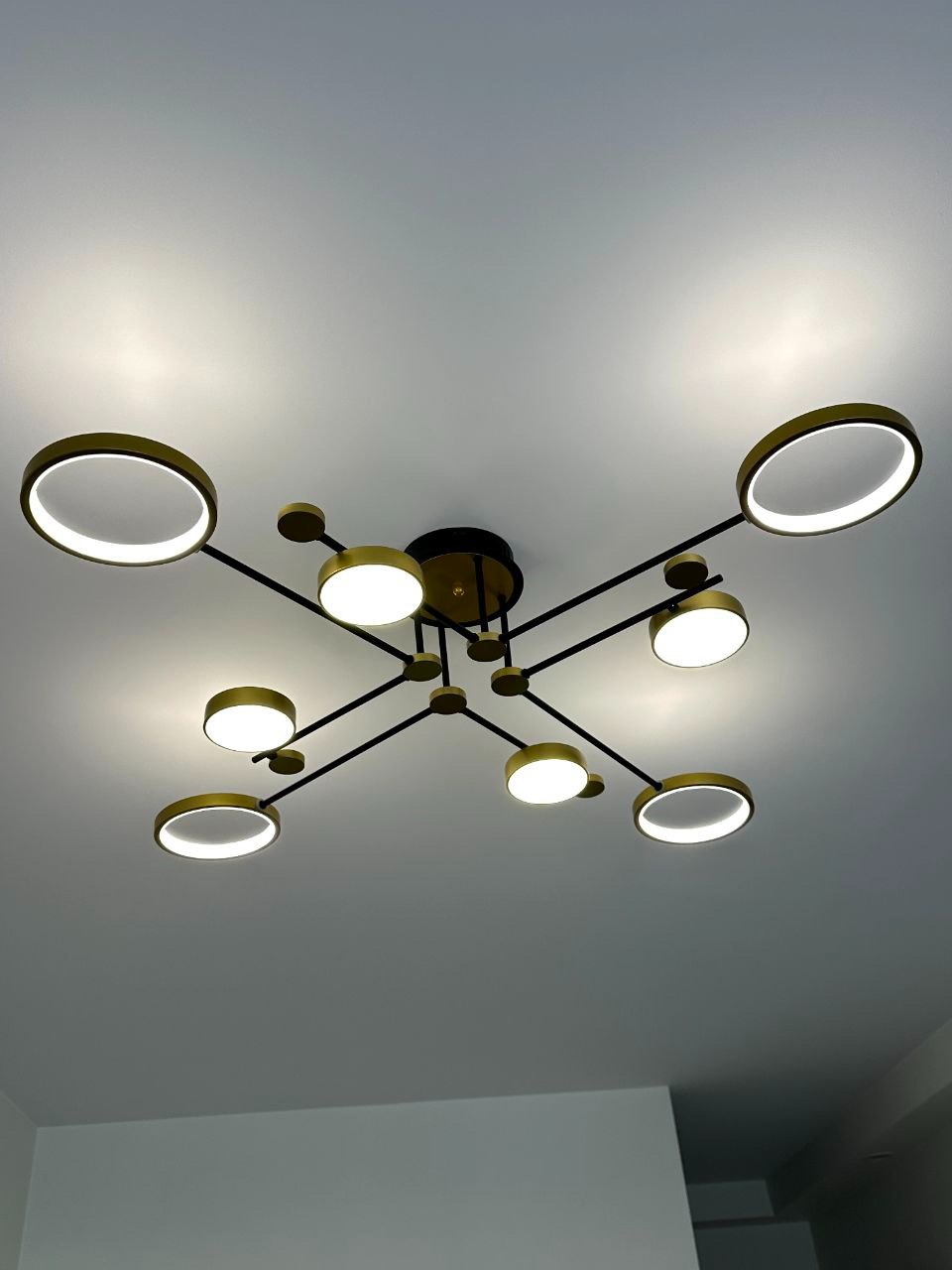 Nowoczesna lampa sufitowa z okrągłymi, złotymi elementami i białym światłem, zamontowana na białym suficie. Czarny stelaż. Ujęcie z dołu.