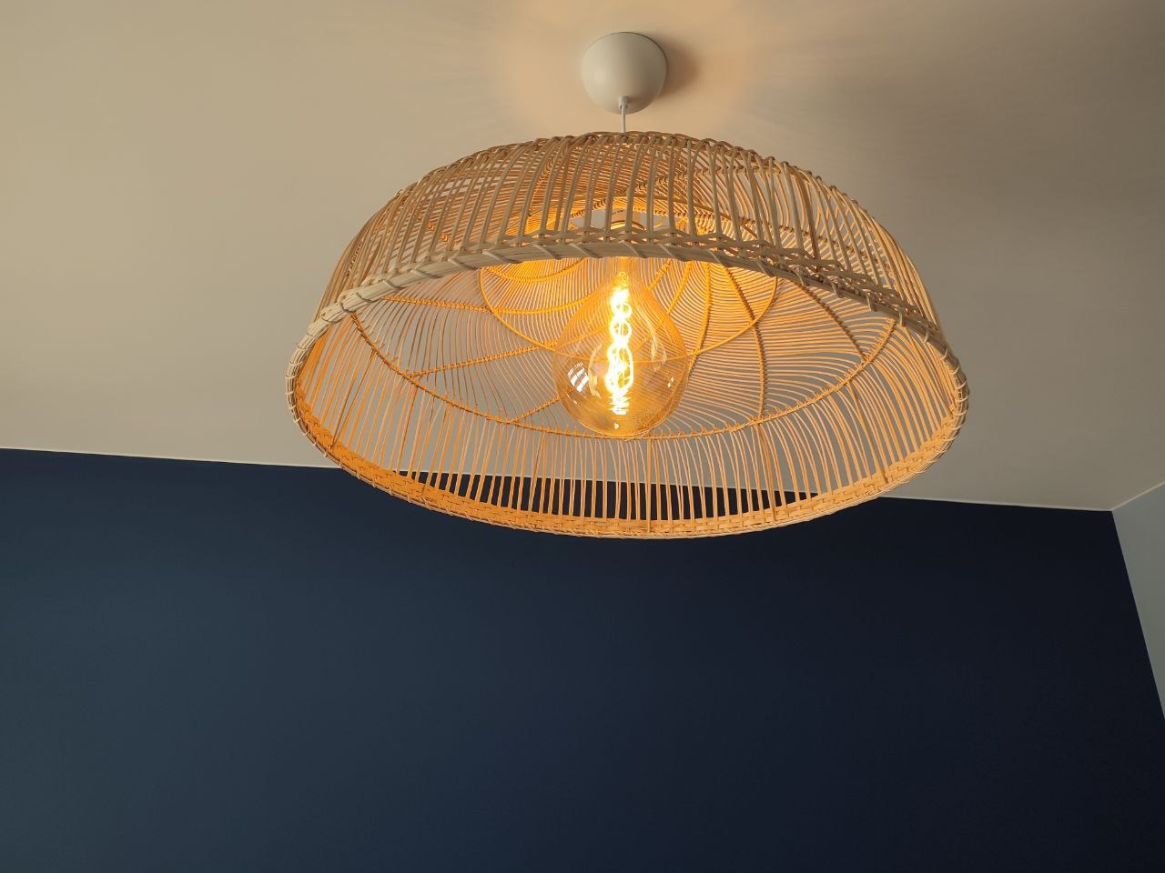Rattanowa lampa wisząca z żarówką Edisona, oświetlająca granatową ścianę. Widok z dołu, ciepłe światło, nowoczesny design.
