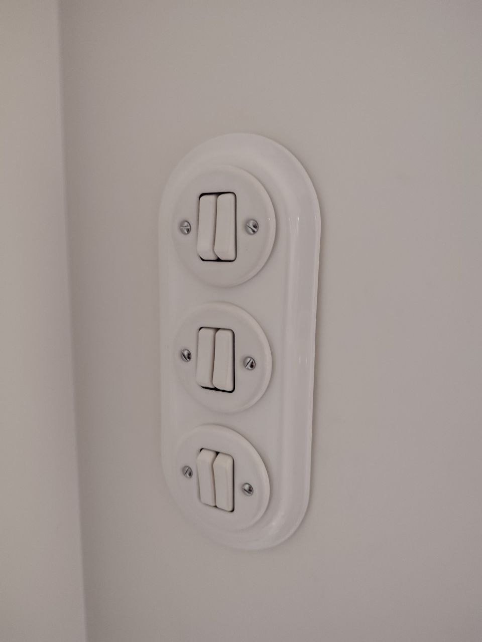 Biały, potrójny włącznik światła w stylu retro, zamontowany na jasnej ścianie. Widoczne śruby mocujące i delikatne cienie podkreślające kształt.