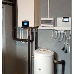 F.E Speed - Biały piec grzewczy Gree z podłączonym zasobnikiem wody, instalacją hydrauliczną i elektryczną na tle szarej ściany. Widoczne rury i elementy sterujące.