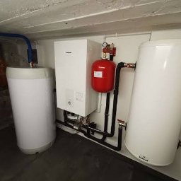 F.E Speed - Biały piec CO, zbiornik wyrównawczy (czerwony) i zasobnik ciepłej wody w kotłowni. Widoczne rury i elementy instalacji grzewczej. Ujęcie w piwnicy.