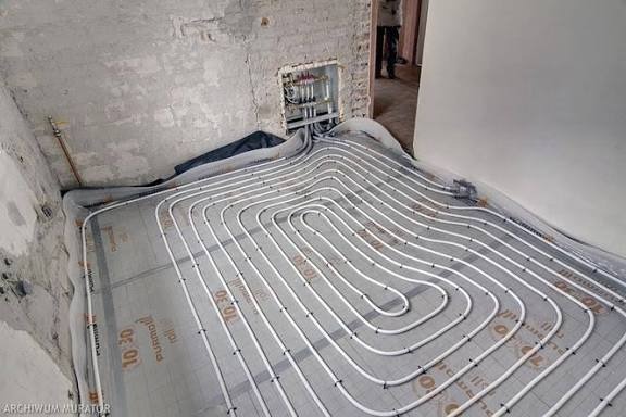 Białe rury ogrzewania podłogowego ułożone spiralnie na szarej macie izolacyjnej w surowym wnętrzu. Widoczne przyłącza do instalacji na ścianie.