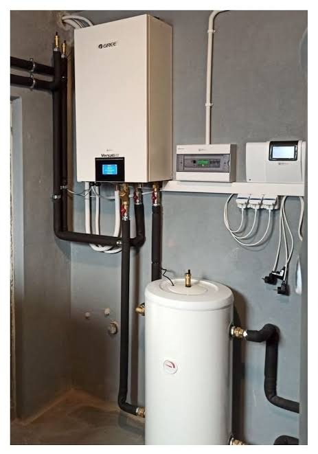 Biały piec grzewczy Gree z podłączonym zasobnikiem wody, instalacją hydrauliczną i elektryczną na tle szarej ściany. Widoczne rury i elementy sterujące.