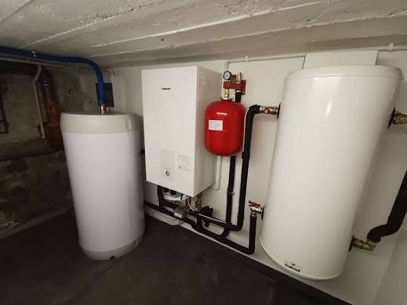 Biały piec CO, zbiornik wyrównawczy (czerwony) i zasobnik ciepłej wody w kotłowni. Widoczne rury i elementy instalacji grzewczej. Ujęcie w piwnicy.