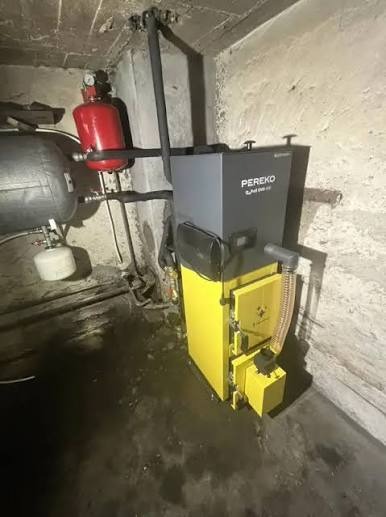 Żółto-szary piec CO marki PEREKO z podłączonymi rurami w kotłowni o surowych ścianach. Widoczny zbiornik wyrównawczy i inne elementy instalacji grzewczej.