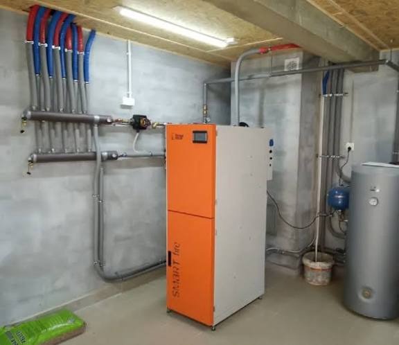 Pomarańczowo-biały piec CO Smart Inc. z podłączonymi rurami instalacji grzewczej na tle szarej ściany w kotłowni. Widoczny zasobnik wody i elementy instalacji.