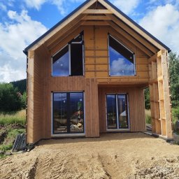 Dom w trakcie budowy z drewnianą elewacją i zamontowanymi oknami. Widoczne elementy konstrukcyjne i izolacyjne. Błękitne niebo odbija się w szybach okien.