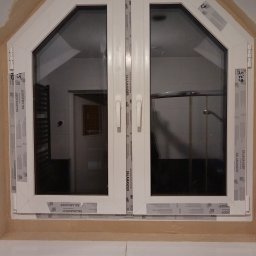 W-Home sp. z o.o. - Dwuskrzydłowe, białe okno PCV o nietypowym, trapezoidalnym kształcie, z widocznymi naklejkami producenta Salamander, zamontowane w surowym otworze okiennym.
