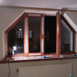 W-Home sp. z o.o. - Montaż okna PCV o nietypowym, trójkątnym kształcie. Widoczne narzędzia pracy i zabezpieczenia. Okno marki Salamander z brązowymi ramami.