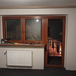 W-Home sp. z o.o. - Brązowe okno z drzwiami balkonowymi, widok na nocne miasto. Na parapecie puszka farby i narzędzia. Biały grzejnik pod oknem. Ujęcie z wnętrza pomieszczenia.