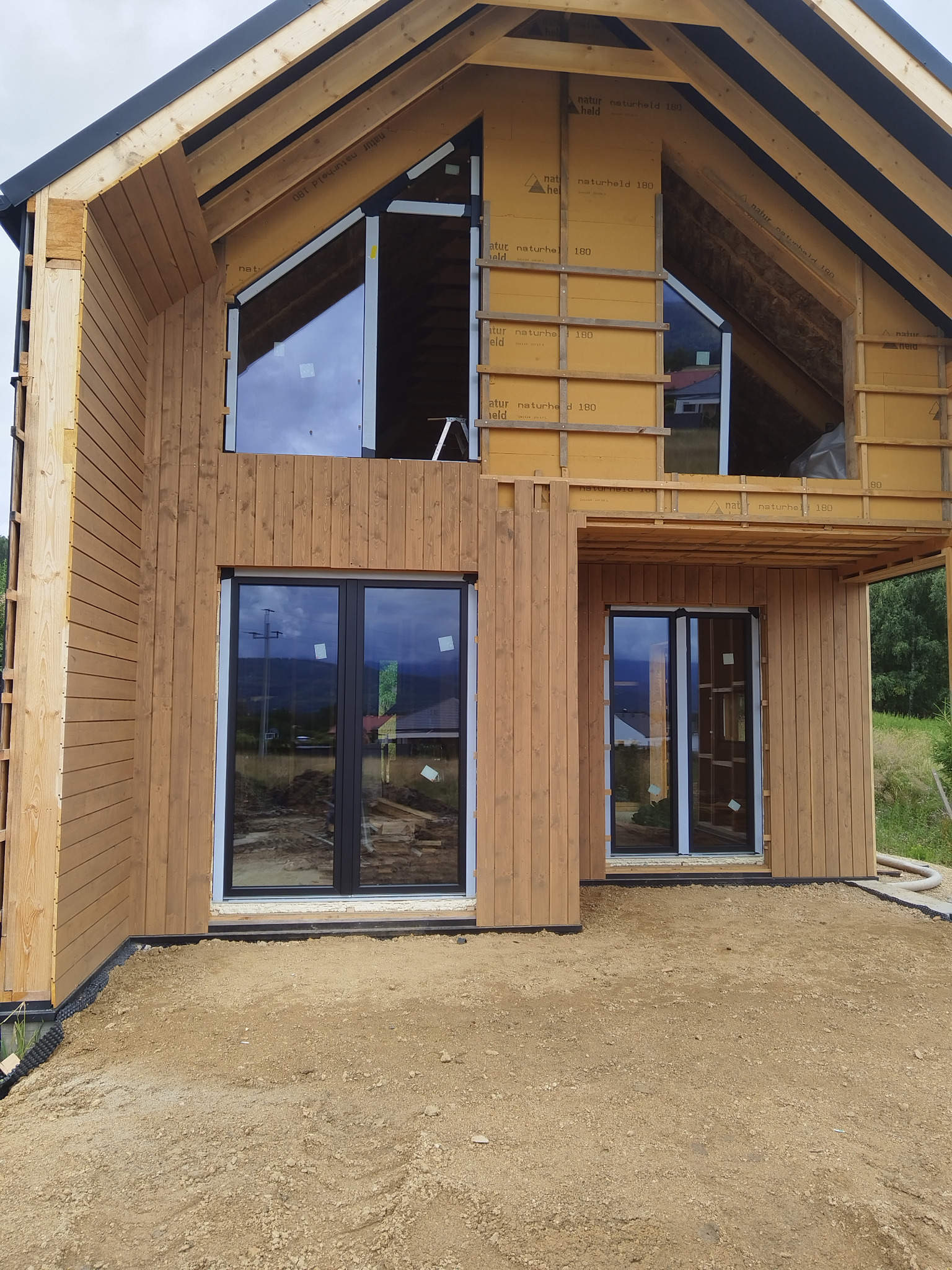 Dom w trakcie budowy z drewnianą elewacją i zamontowanymi oknami. Widoczne elementy konstrukcyjne i izolacyjne. Okna z czarnymi ramami.