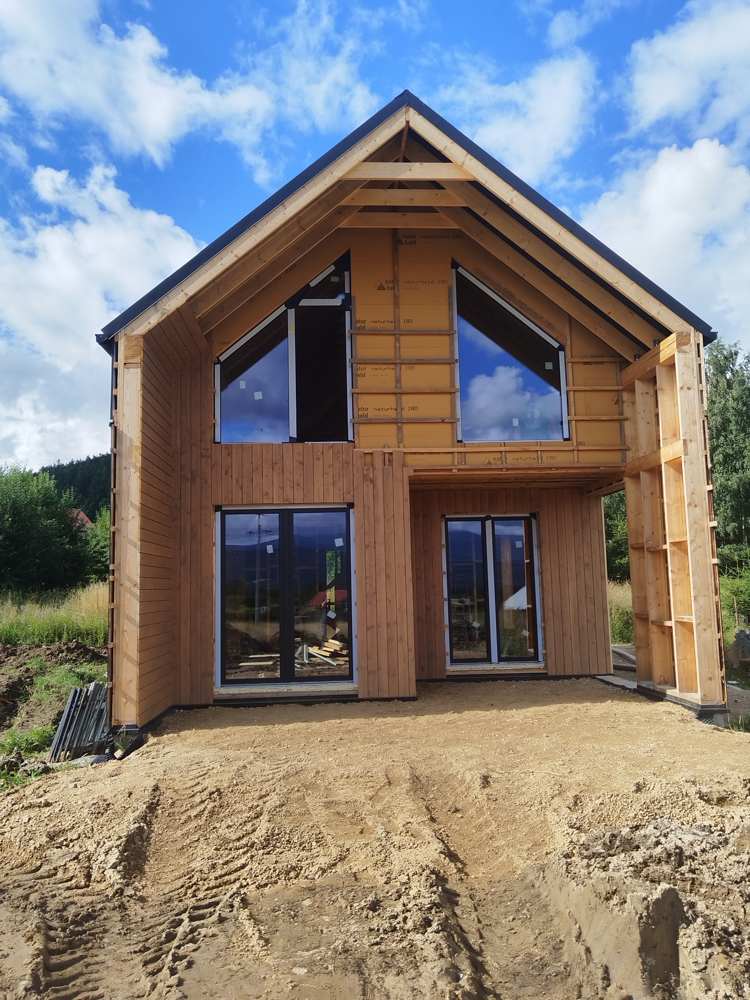 Dom w trakcie budowy z drewnianą elewacją i zamontowanymi oknami. Widoczne elementy konstrukcyjne i izolacyjne. Błękitne niebo odbija się w szybach okien.