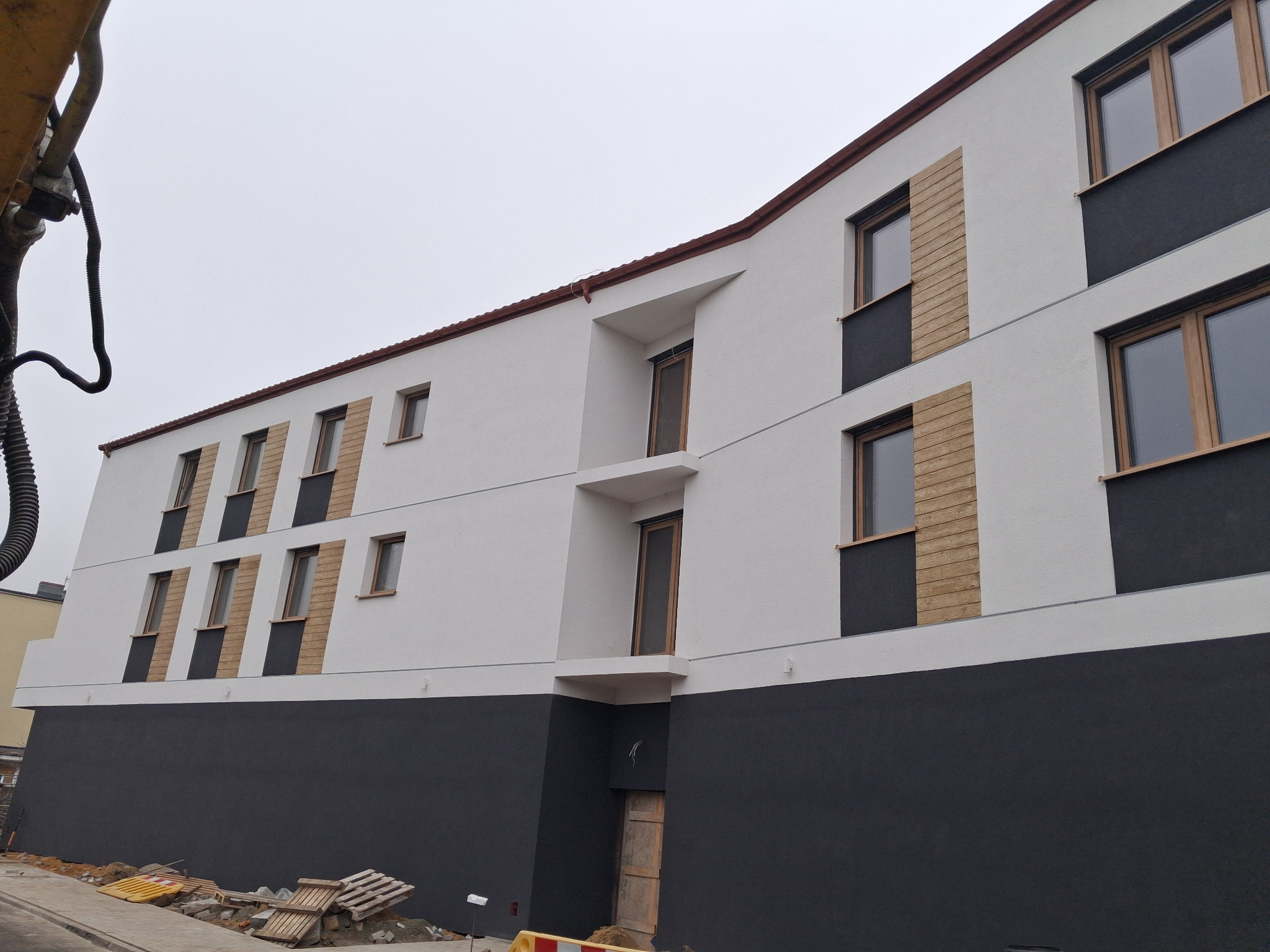 Nowoczesny budynek wielorodzinny z oknami w drewnianych ramach i elementami dekoracyjnymi. Widoczne rusztowania i materiały budowlane.