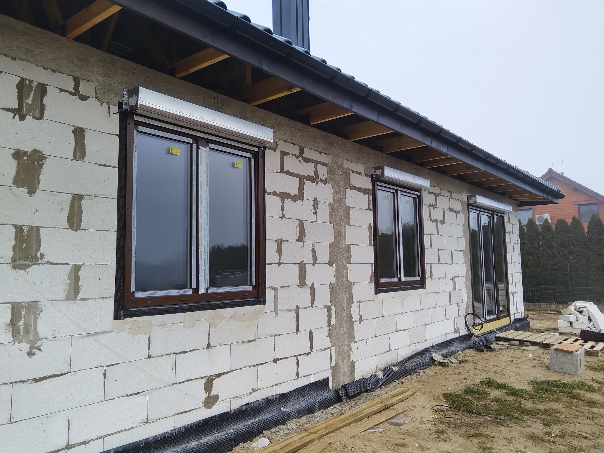 Surowy budynek z brązowymi oknami i zamontowanymi roletami zewnętrznymi w kolorze srebrnym. Widoczne bloczki betonu komórkowego i elementy konstrukcji dachu.