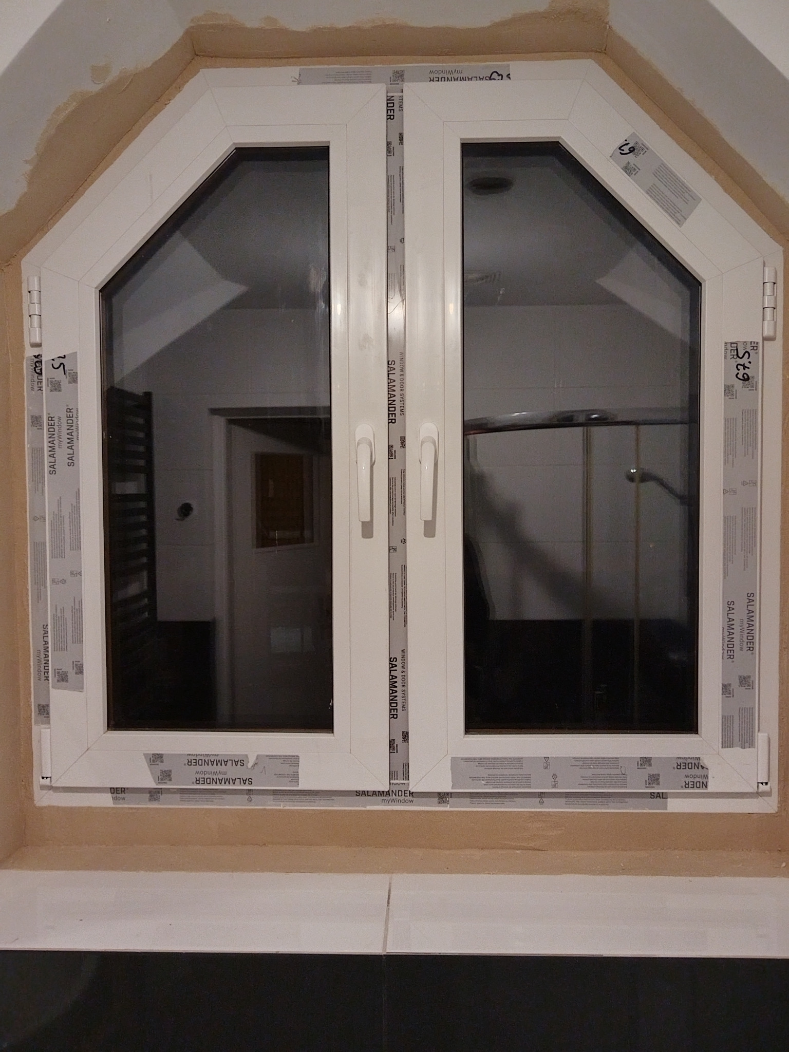 Dwuskrzydłowe, białe okno PCV o nietypowym, trapezoidalnym kształcie, z widocznymi naklejkami producenta Salamander, zamontowane w surowym otworze okiennym.