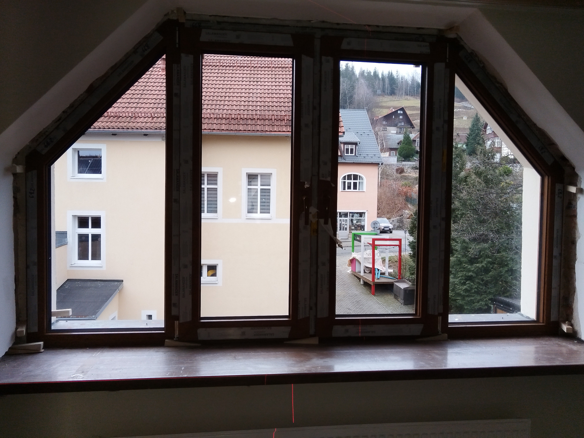 Nietypowe okno z brązowymi ramami w kształcie trapezu, widok na budynki i drzewa. Widoczne elementy montażowe i laserowy wskaźnik poziomu.