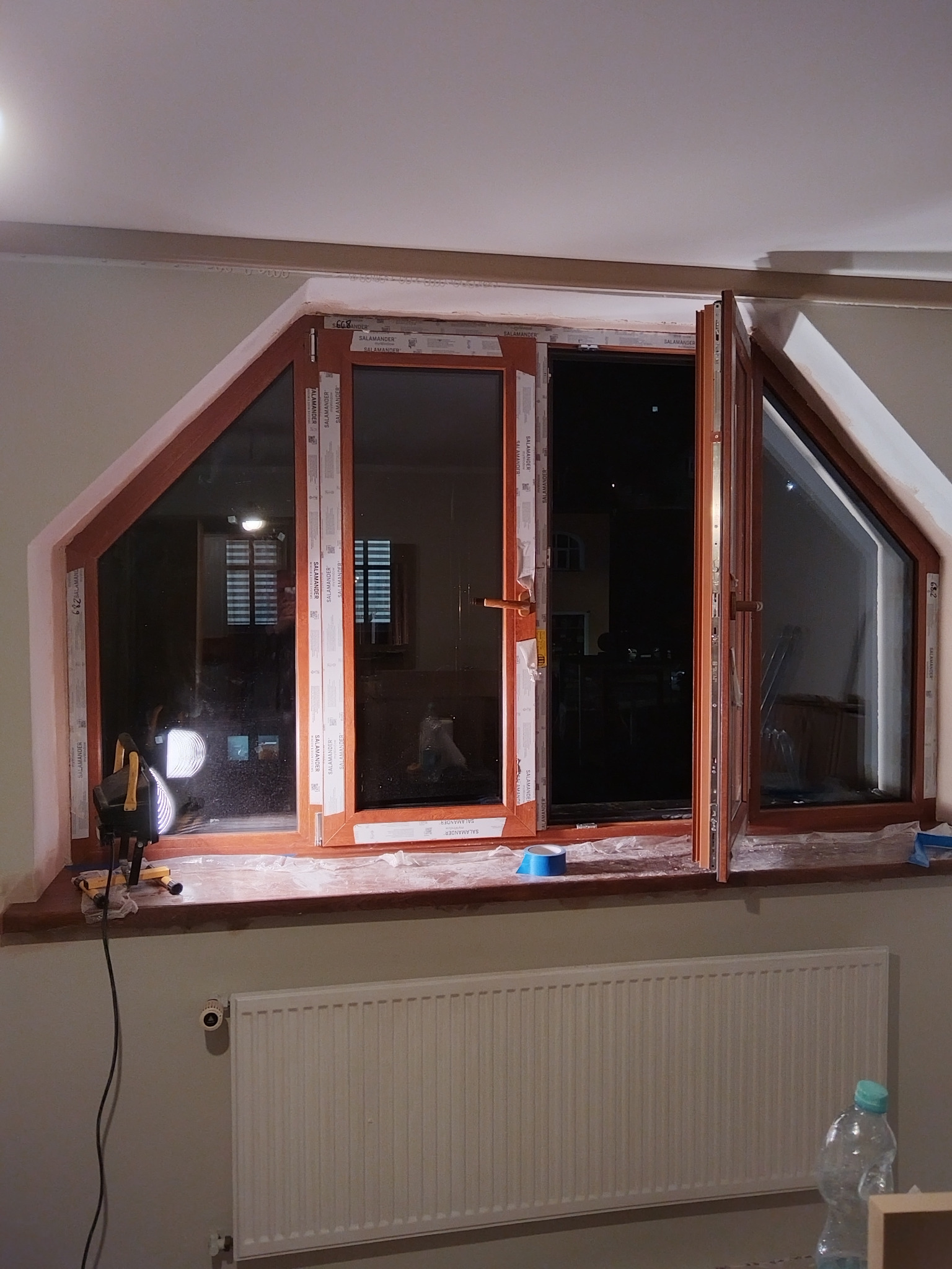 Montaż okna PCV o nietypowym, trójkątnym kształcie. Widoczne narzędzia pracy i zabezpieczenia. Okno marki Salamander z brązowymi ramami.