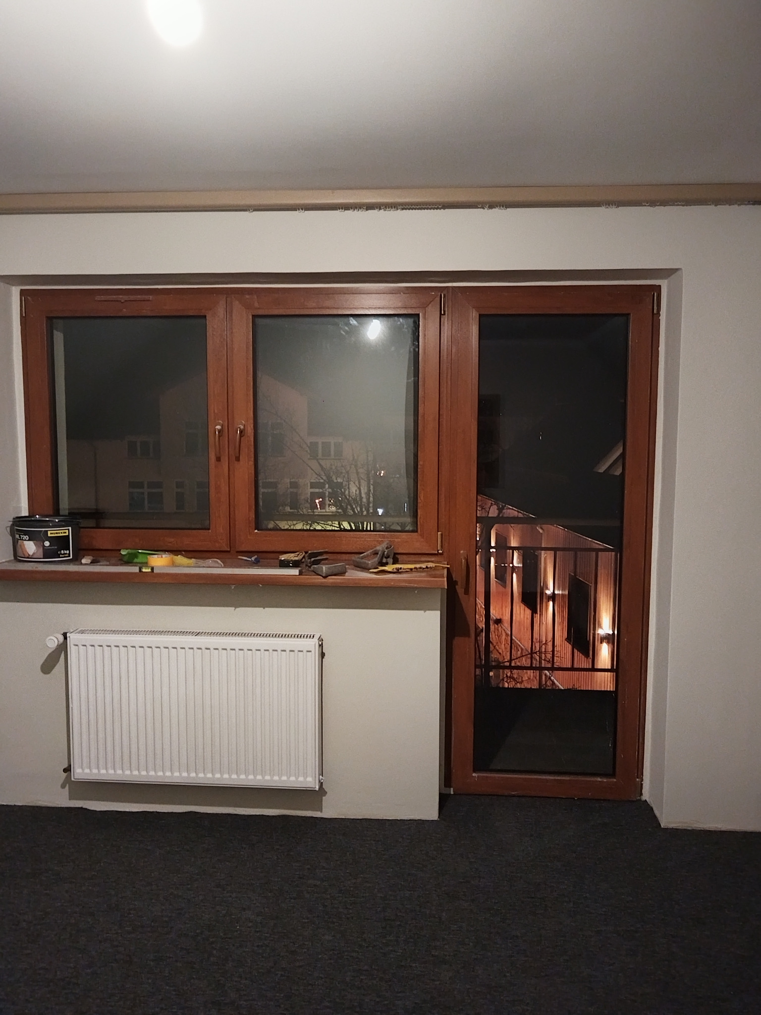 Brązowe okno z drzwiami balkonowymi, widok na nocne miasto. Na parapecie puszka farby i narzędzia. Biały grzejnik pod oknem. Ujęcie z wnętrza pomieszczenia.