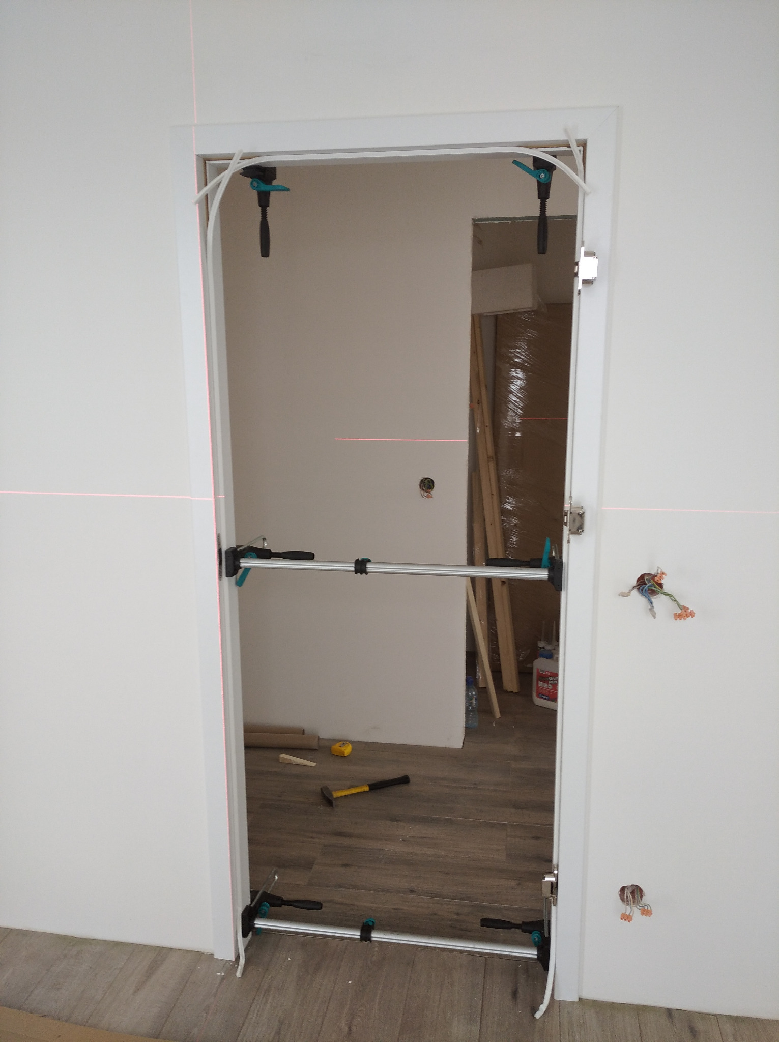 Montaż ościeżnicy drzwiowej z użyciem rozpórki i lasera krzyżowego. Widoczne narzędzia i elementy instalacji elektrycznej. Podłoga wyłożona panelami.