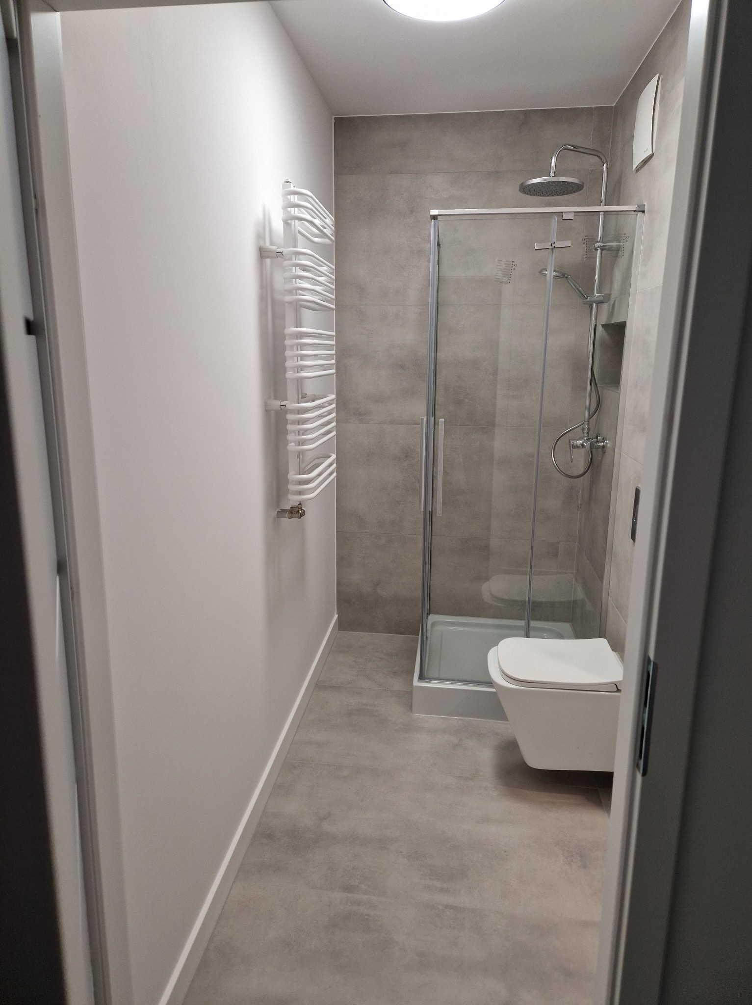 Nowoczesna łazienka po remoncie: szara płytka, kabina prysznicowa, podwieszana toaleta i biały grzejnik drabinkowy. Minimalistyczny design, neutralna kolorystyka.
