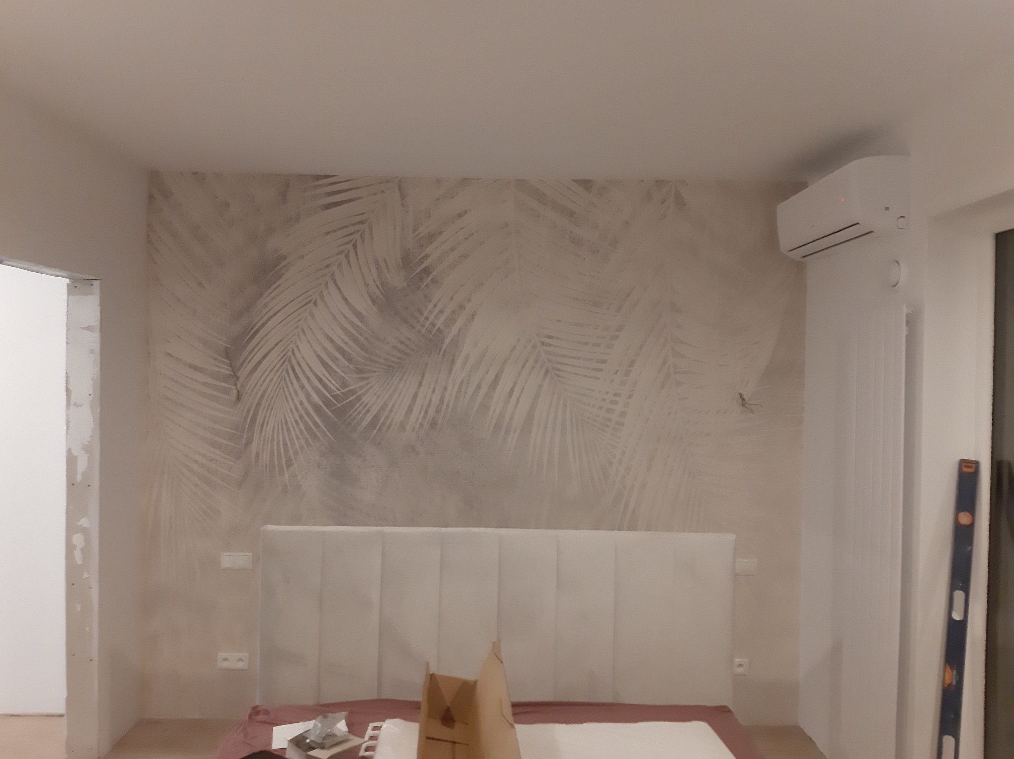 Pokój po malowaniu z motywem liści palmowych na ścianie za wezgłowiem łóżka. Widoczny klimatyzator i poziomica oparta o ścianę. Częściowo widoczne narzędzia.