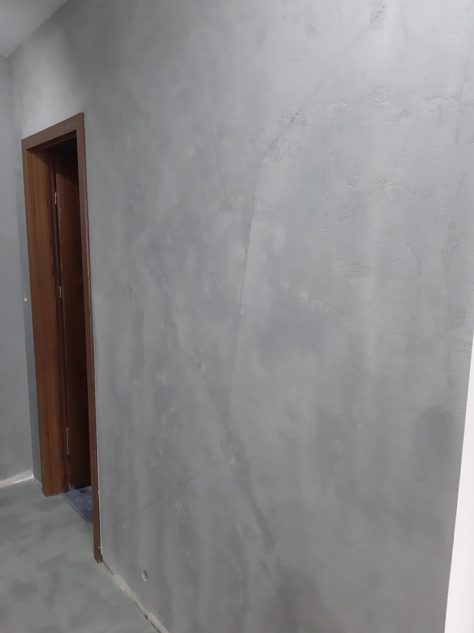 Szara ściana z efektem dekoracyjnym i drewniana ościeżnica drzwiowa. Minimalistyczne wykończenie wnętrza, widoczne detale faktury na ścianie.