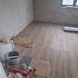 Ukończona podłoga z płytek imitujących drewno w pokoju. Widoczne narzędzia glazurnicze i resztki kartonów po płytkach. Ściany przygotowane do malowania.