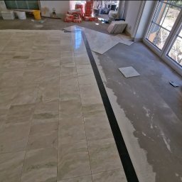 Układanie beżowych płytek podłogowych z czarnym pasem dekoracyjnym. Częściowo ułożona podłoga, widoczne worki z klejem i narzędzia glazurnicze.