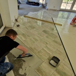 Usługi Budowlane Paweł Łozowski - Układanie płytek podłogowych w jasnym wnętrzu. Widoczny glazurnik rozprowadzający klej, poziomica i krzyżyki dystansowe. Duże okna w tle.