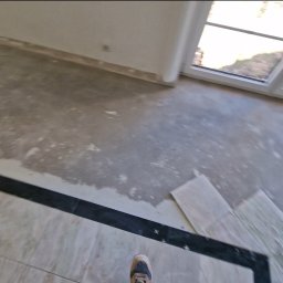 Usługi Budowlane Paweł Łozowski - Wnętrze w trakcie prac wykończeniowych: częściowo ułożona podłoga z płytek, widoczne fragmenty wylewki. W tle okno i ściany przygotowane do malowania.