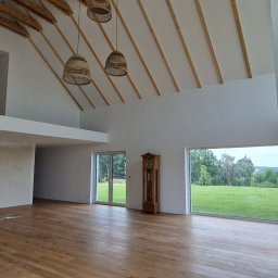 Usługi Budowlane Paweł Łozowski - Jasny parkiet w przestronnym salonie z drewnianym stropem i widokiem na zieleń. Trzy wiklinowe lampy wiszą nad podłogą. Klasyczny zegar stoi przy oknie.