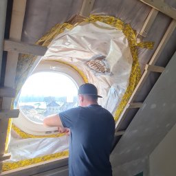 Usługi Budowlane Paweł Łozowski - Montaż okna okrągłego na poddaszu. Widoczna izolacja folią paroizolacyjną. Fachowiec w trakcie pracy, drewniana konstrukcja dachu, grzejnik i narzędzia w pomieszczeniu.
