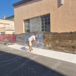 Usługi Budowlane Paweł Łozowski - Pracownik w trakcie malowania elewacji budynku natryskowo, z użyciem agregatu malarskiego. Część ściany już pomalowana na biało, widoczne zabezpieczenia podłoża.