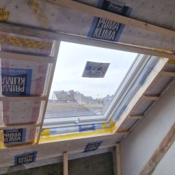 Usługi Budowlane Paweł Łozowski - Montaż okna dachowego w skośnym dachu, widoczne drewniane ramy, folia paroizolacyjna z nadrukiem, fragment poddasza w trakcie prac wykończeniowych.