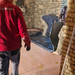 Usługi Budowlane Paweł Łozowski - Prace izolacyjne w piwnicy ze sklepieniem łukowym: ekipa montuje czarną matę izolacyjną na różowej warstwie styropianu, widoczne żółte elementy mocujące.