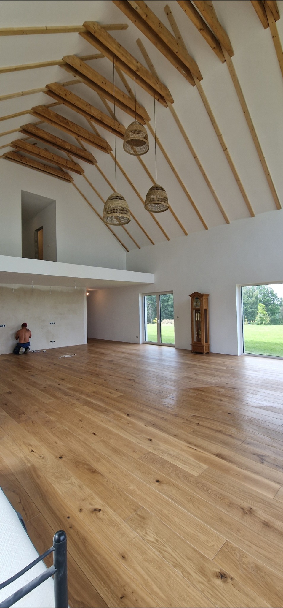 Jasna, przestronna sala z drewnianą podłogą i widoczną konstrukcją dachu. Pracownik szpachluje ścianę. W tle okna z widokiem na zieleń i wysoki zegar.