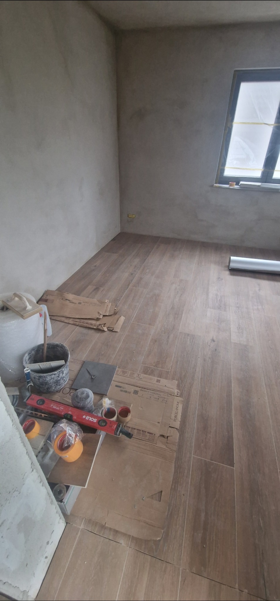 Ukończona podłoga z płytek imitujących drewno w pokoju. Widoczne narzędzia glazurnicze i resztki kartonów po płytkach. Ściany przygotowane do malowania.