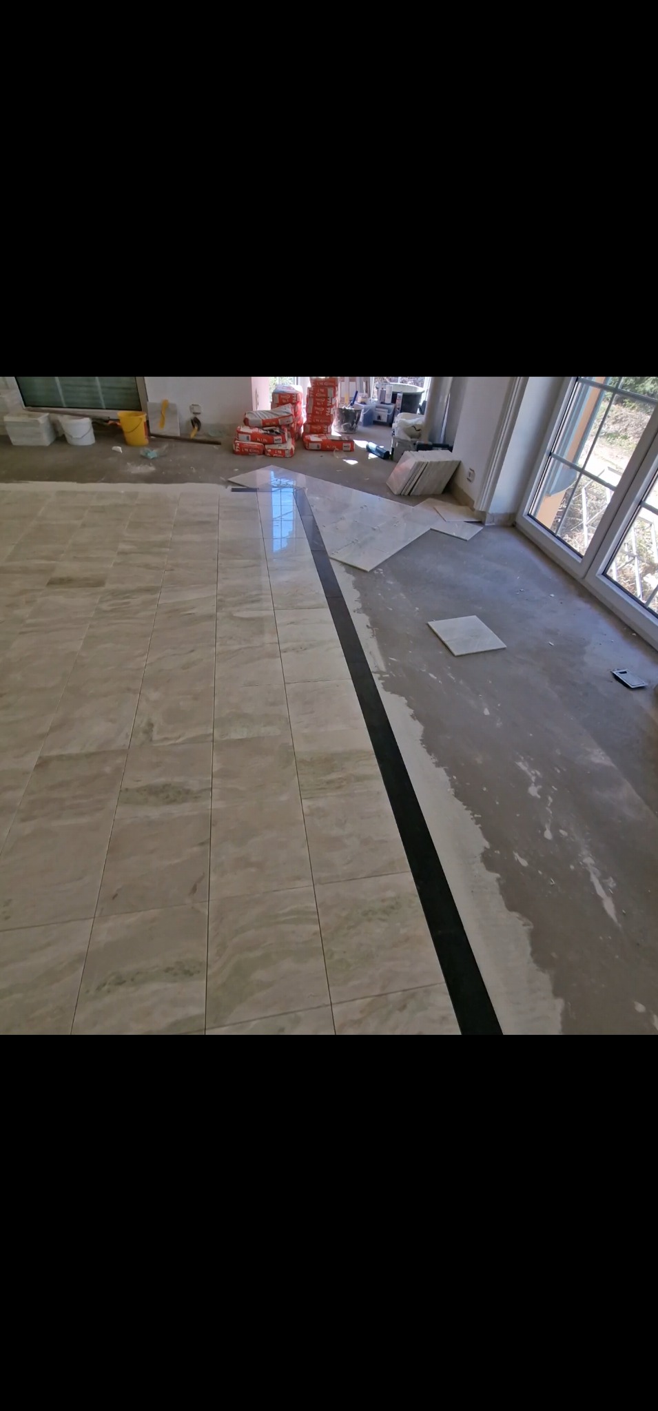 Układanie beżowych płytek podłogowych z czarnym pasem dekoracyjnym. Częściowo ułożona podłoga, widoczne worki z klejem i narzędzia glazurnicze.