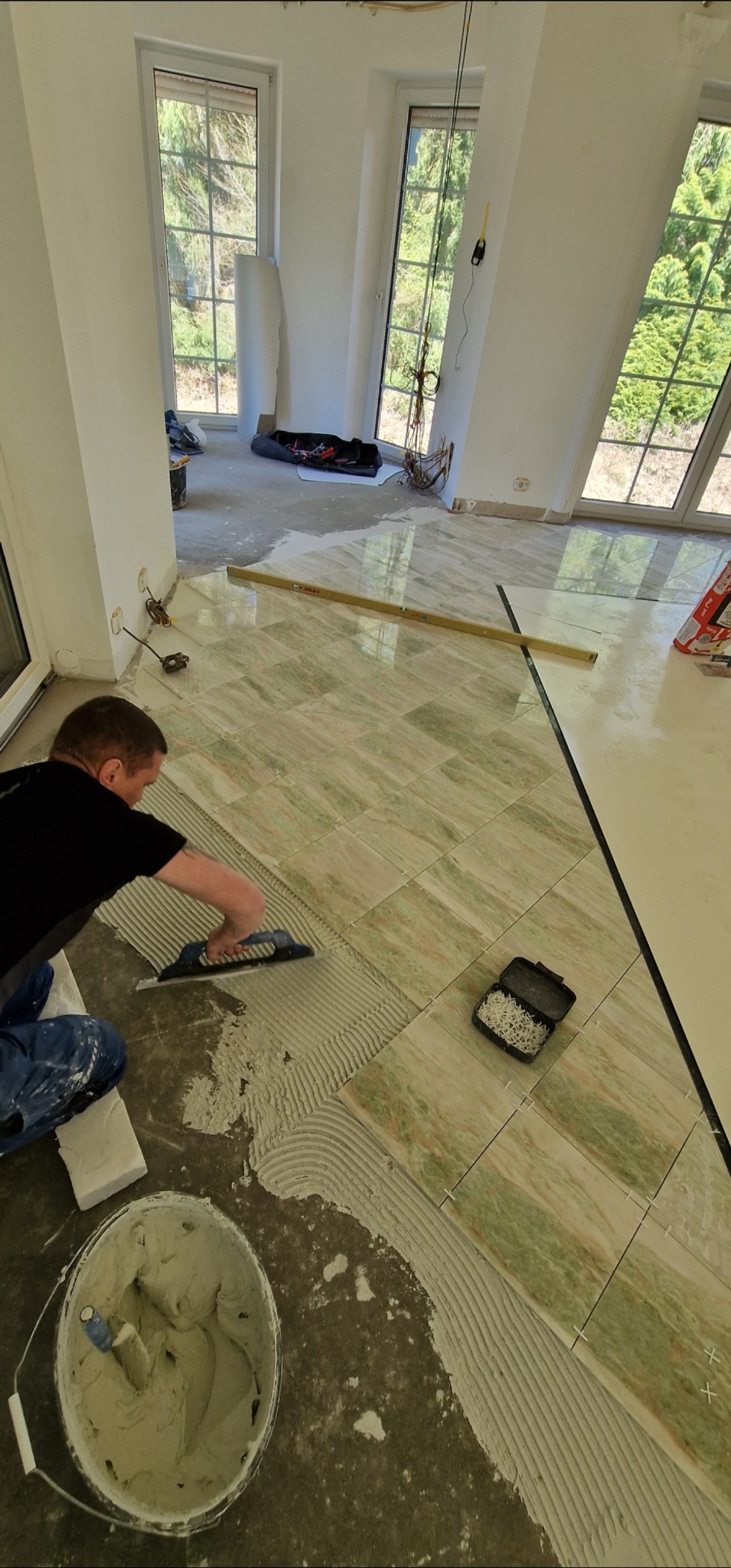 Układanie płytek podłogowych w jasnym wnętrzu. Widoczny glazurnik rozprowadzający klej, poziomica i krzyżyki dystansowe. Duże okna w tle.