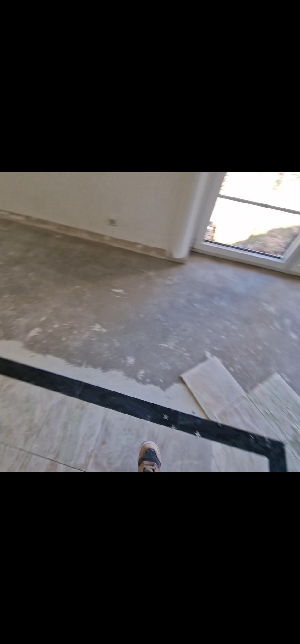 Wnętrze w trakcie prac wykończeniowych: częściowo ułożona podłoga z płytek, widoczne fragmenty wylewki. W tle okno i ściany przygotowane do malowania.