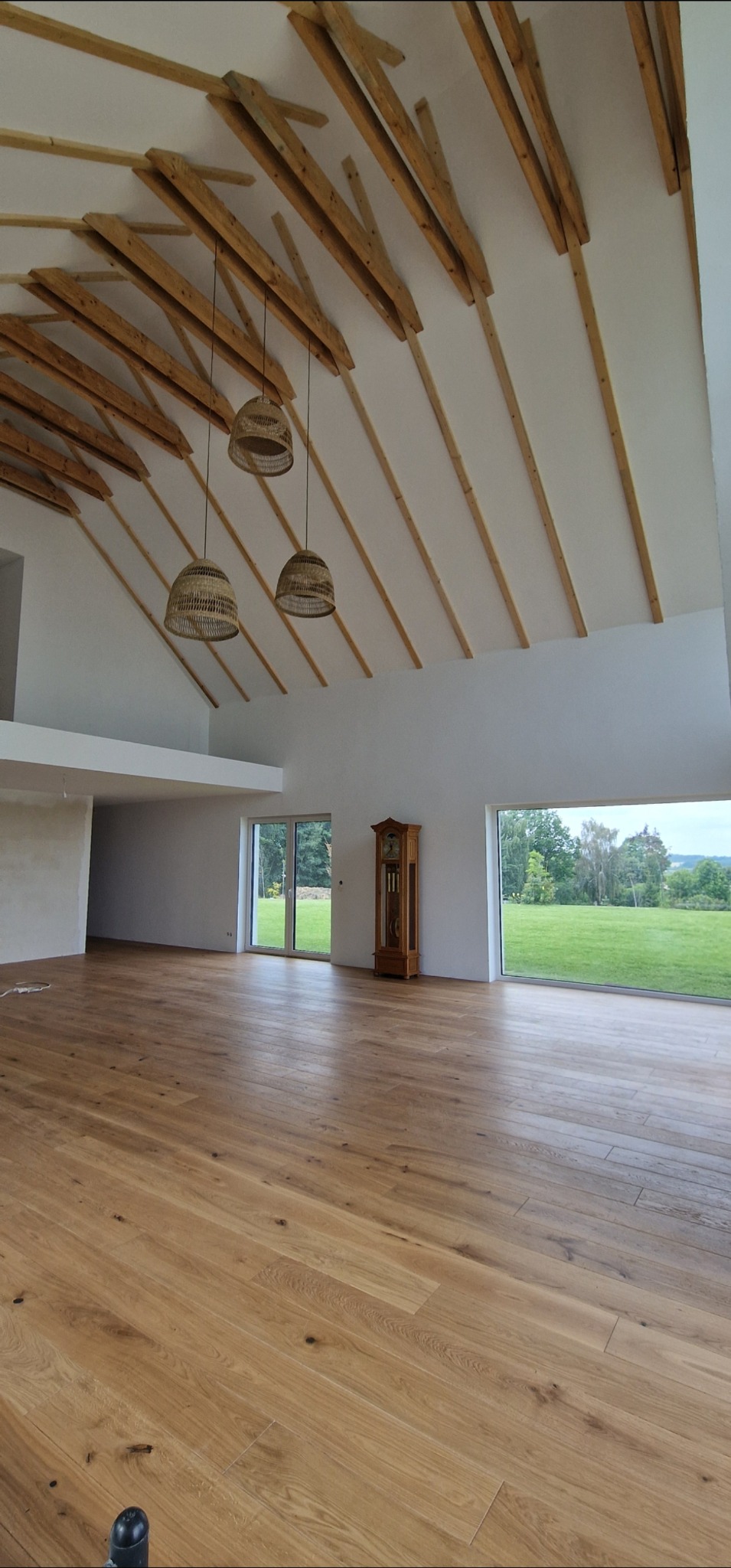 Jasny parkiet w przestronnym salonie z drewnianym stropem i widokiem na zieleń. Trzy wiklinowe lampy wiszą nad podłogą. Klasyczny zegar stoi przy oknie.