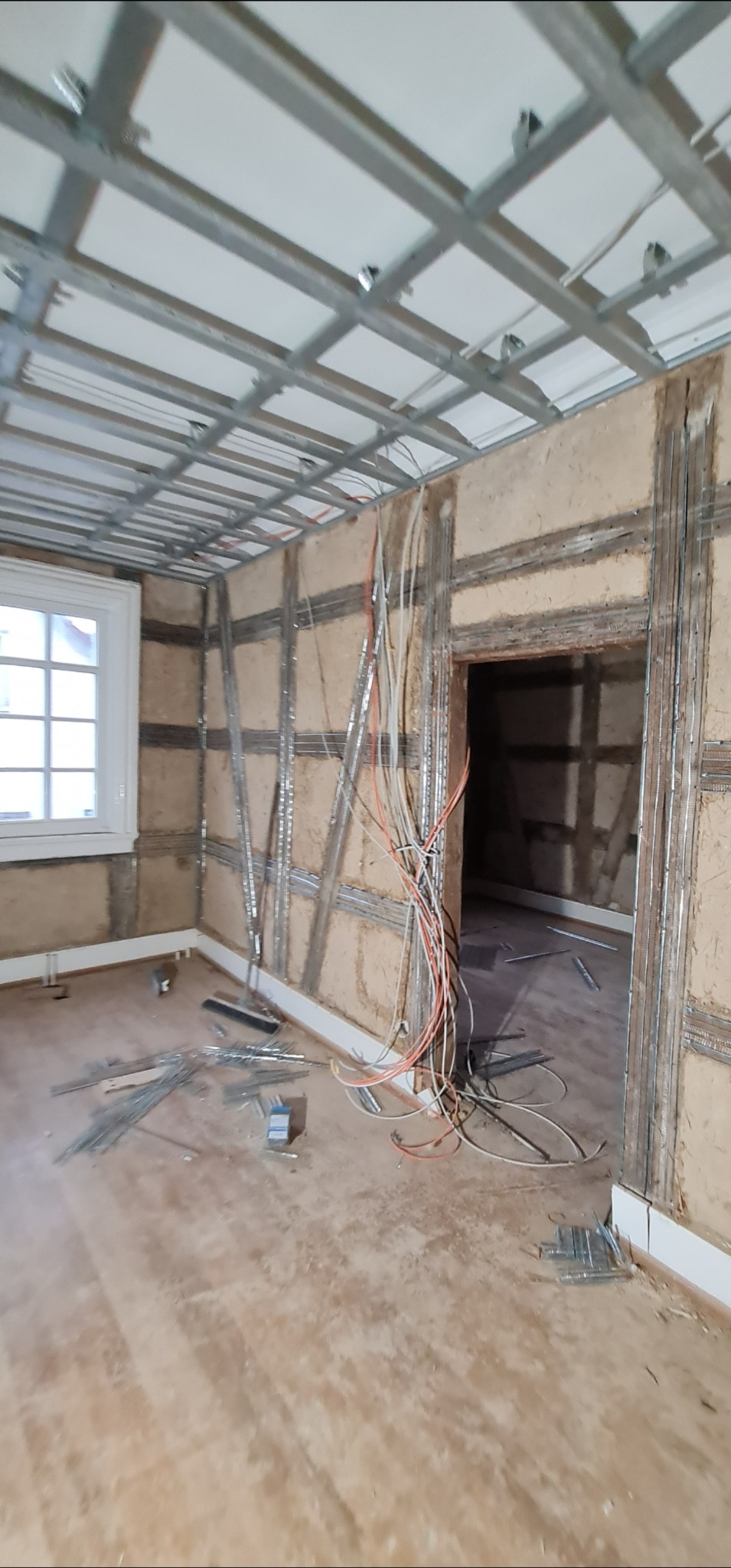 Pokój w trakcie montażu stelaża sufitu podwieszanego i ścian działowych z profili metalowych. Widoczne przewody elektryczne i okno z białymi ramami.