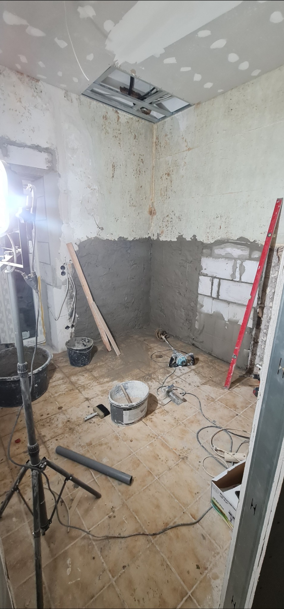 Wnętrze w trakcie prac remontowych: częściowo otynkowana ściana, widoczne narzędzia, poziomica, wiadro z zaprawą, lampa na statywie. Sufit z elementami konstrukcji pod płytę gipsową.