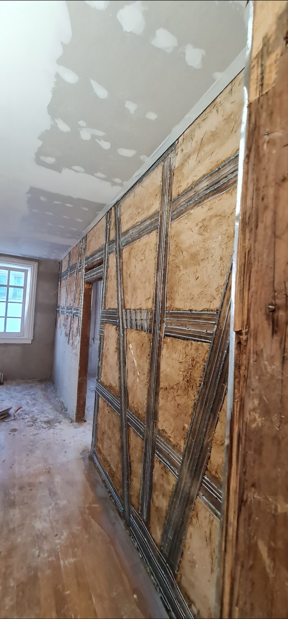 Ściana z widoczną konstrukcją nośną i wypełnieniem z gliny, przygotowana do dalszych prac wykończeniowych. Sufit częściowo pokryty gładzią. Widoczne okno i fragment podłogi.