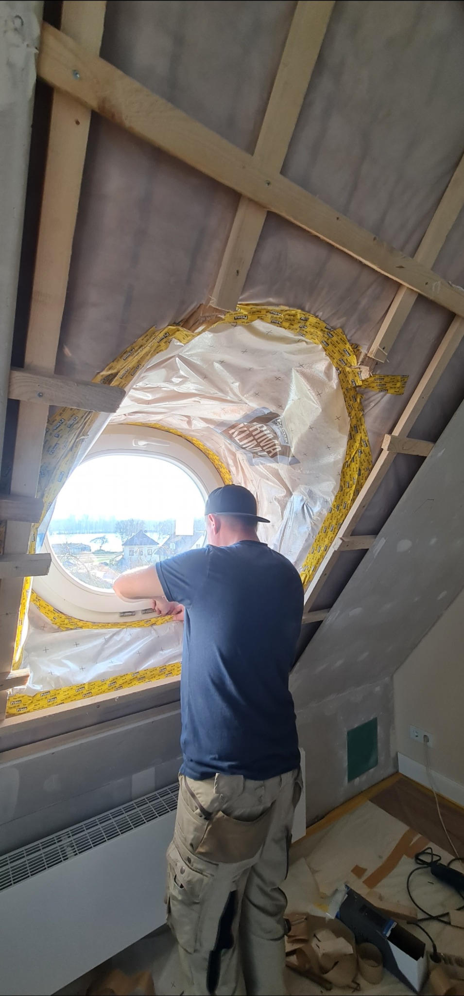 Montaż okna okrągłego na poddaszu. Widoczna izolacja folią paroizolacyjną. Fachowiec w trakcie pracy, drewniana konstrukcja dachu, grzejnik i narzędzia w pomieszczeniu.