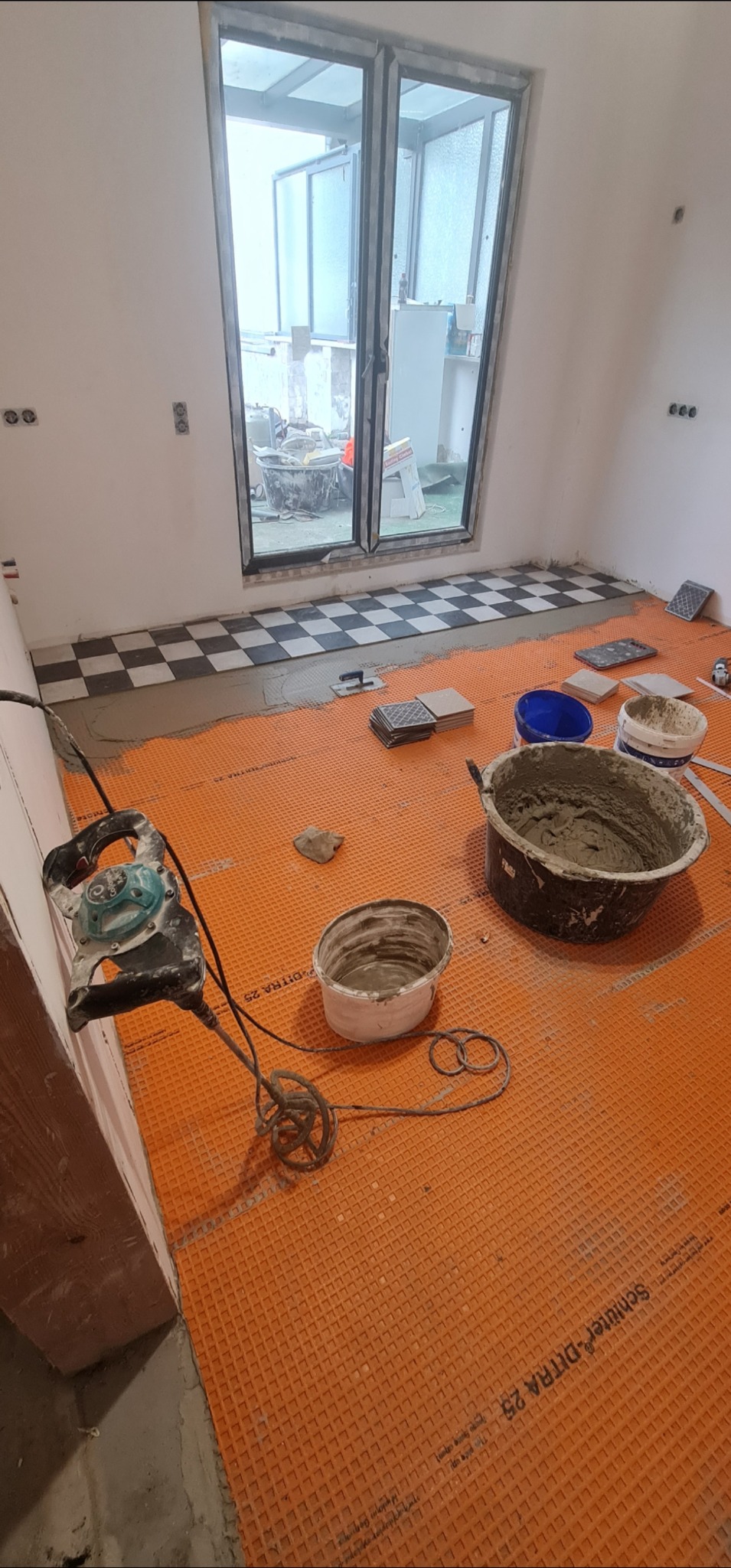 Układanie płytek podłogowych: widoczna pomarańczowa mata izolacyjna, częściowo ułożona szachownica, narzędzia glazurnicze i zaprawa. Prace w toku, widok na balkon.