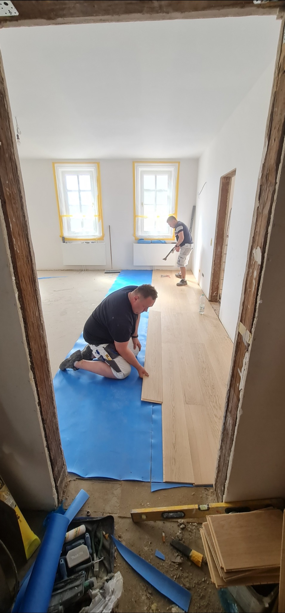 Układanie jasnego parkietu w pokoju z białymi ścianami. Dwóch mężczyzn montuje panele na niebieskiej folii. Widok zza drewnianej ramy drzwi.
