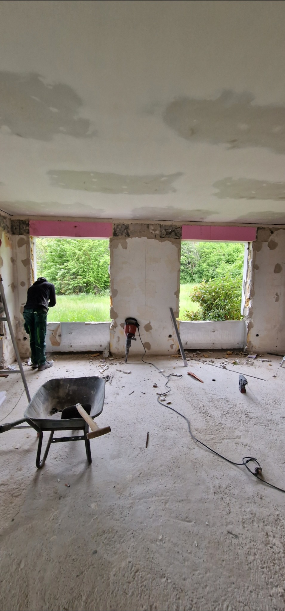 Wnętrze w trakcie demontażu: pracownik w zielonym kombinezonie, taczka, młot udarowy Hiltti, odsłonięte okna z widokiem na zieleń. Etap przygotowawczy do remontu.