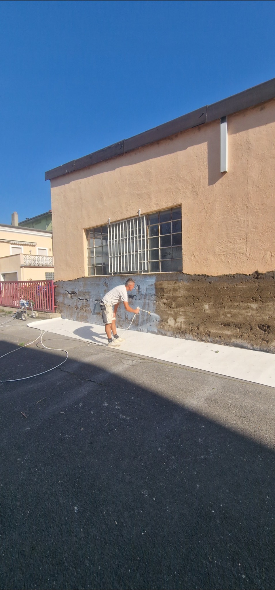 Pracownik w trakcie malowania elewacji budynku natryskowo, z użyciem agregatu malarskiego. Część ściany już pomalowana na biało, widoczne zabezpieczenia podłoża.